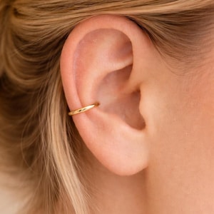 May include: Close-up of a gold ear cuff. The cuff is a simple, smooth band that curves around the ear cartilage. The skin tone is fair, and the hair is blonde. The jewelry is a minimalist accessory.