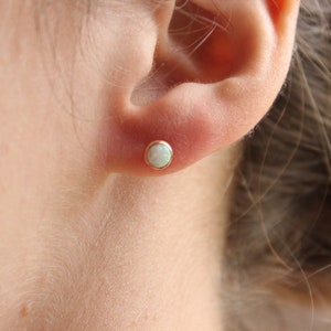 May include: A close-up of a person's ear with a small, round, white opal stud earring in a gold setting.