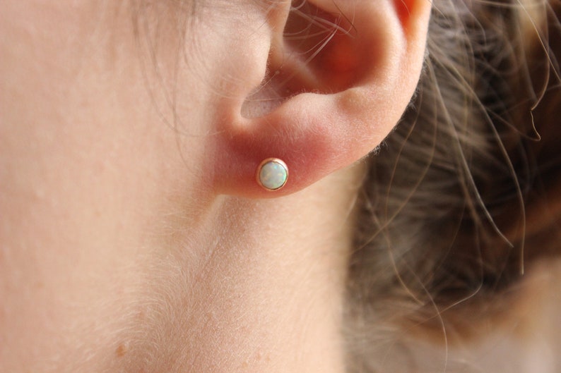 May include: A close-up of a person's ear with a small, round, white opal stud earring in a gold setting.