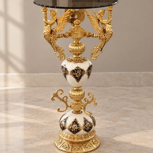 Mesita auxiliar blanca, azul y dorada con tapa de cristal ornamentada y figuras aladas.