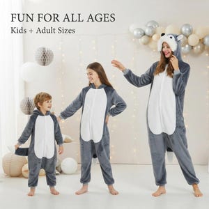 May include: A grey and white wolf-themed onesie featuring a hood with ears and a tail. The text FUN FOR ALL AGES Kids + Adult Sizes is visible. The garment is made of soft plush fabric with a front zip closure, long sleeves, and cuffed ankles.