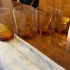 May include: A set of four amber-colored glass tumblers, each featuring a cylindrical shape with a series of horizontal ridges encircling the lower portion of the glass, providing a textured grip and a distinct visual detail.