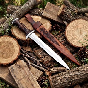 May include: A double-edged dagger with a polished silver blade and a dark brown handle featuring a woven pattern. The dagger includes a matching brown wooden sheath engraved with intricate knotwork designs and a leather strap for carrying.