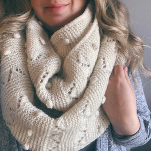 May include: A white knitted infinity scarf with a textured pattern and small white pom-poms.