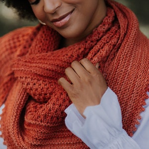 May include: A close-up of a woman wearing a rust-colored knitted shawl with tassels. The shawl is crocheted with a delicate stitch pattern. The woman is wearing a white blouse with ruffled sleeves.