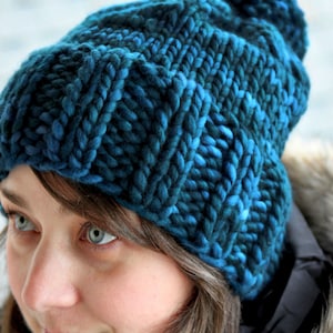 May include: A blue and green knitted beanie hat with a pompom on top. The hat is made with thick yarn and has a chunky knit texture.