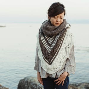 May include: A woman wearing a brown and white knitted shawl with a lacy pattern. She is standing on a rocky beach with the ocean in the background.