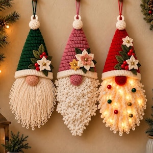Puede incluir: Tres adornos colgantes de gnomos hechos de ganchillo. El primero tiene un gorro verde oscuro, el segundo uno rosa empolvado y el tercero uno rojo con luces integradas. Cada uno presenta una barba de color crema y detalles florales.