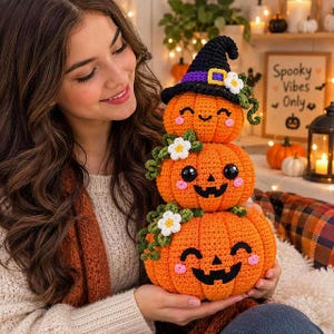 Può includere: Una pila di tre zucche all'uncinetto arancioni con facce in feltro nero e guance rosa. La zucca in cima indossa un cappello da strega nero con una fascia viola e un fiore bianco. Viticci verdi e fiori bianchi decorano la pila di zucche.