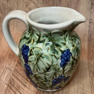 May include: A ceramic pitcher with a white handle and spout. The body is decorated with green leaves and blue grape clusters. The pitcher has a bulbous shape and a wide opening.