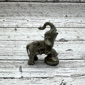 May include: A gray metal elephant figurine with its trunk raised, standing on a round base. The elephant is decorated with a patterned design.