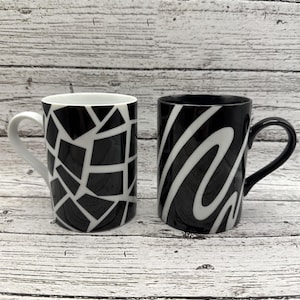 May include: Two ceramic mugs with black and white designs. One mug features a geometric pattern of black shapes on a white background. The other mug has a black base with white, wavy stripes. Both mugs have a curved handle.