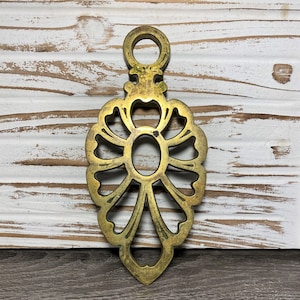 May include: A gold-toned metal trivet with a decorative floral design. The trivet has a loop at the top for hanging.