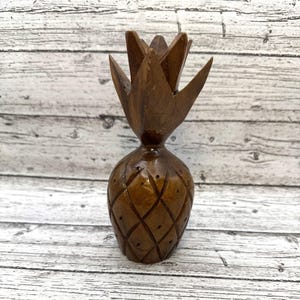 May include: A handcrafted wooden pineapple figurine. The brown pineapple has a detailed diamond pattern on the body and a spiky crown. The figurine is displayed on a light-colored, wooden surface.