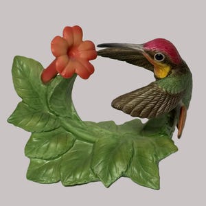 May include: A ceramic figurine featuring a hummingbird with a red crown, yellow throat, and green wings, hovering near a bright orange trumpet-shaped flower. The bird and flower are attached to a base of sculpted green leaves and stems.