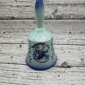May include: A ceramic bell with a light blue and dark blue gradient handle and body. The bell features a cartoon illustration and the text "Dogpatch U.S.A." and "Gifts Capp Enterprises, Inc."