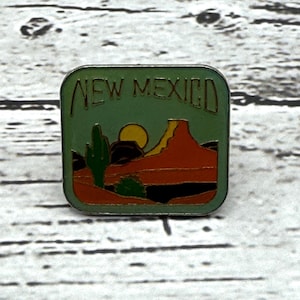 May include: A square pin featuring a New Mexico landscape design. The pin has a green background with the words "NEW MEXICO" at the top. The design includes a desert scene with a cactus, mountains, and a sun.