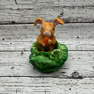 May include: A ceramic figurine of a brown rabbit with long ears, holding a small orange carrot with green leaves. The rabbit is positioned inside a sculpted green cabbage head with detailed, layered leaves forming a base for the animal figure.