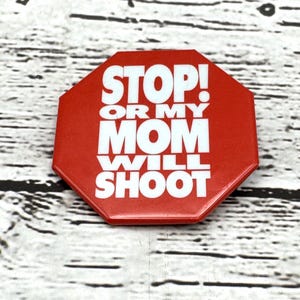 May include: A red, octagonal button with the words "STOP! OR MY MOM WILL SHOOT" in white block letters. The button is set against a light wood grain background.