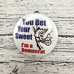 May include: A white political button with the text "You Bet Your Sweet I'm a Democrat" in blue and red. A black and white illustration of a donkey is on the right side of the button.