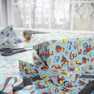May include: Light blue wrapping paper featuring a repeating pattern of brown monkeys, yellow hats, red and blue balloons, bicycles, books, ice cream cones, and cars. The paper is shown wrapped around rectangular boxes with black scissors nearby.