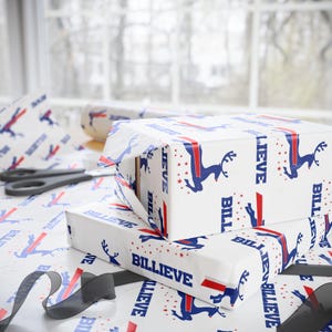 May include: White wrapping paper featuring a repeating pattern of blue reindeer silhouettes, red stars, and the word BILLIEVE in bold blue capital letters. The paper is shown covering two rectangular boxes, with a roll and scissors nearby.