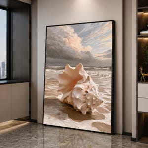 Op de afbeelding: Een ingelijste verticale wandkunstprint met een grote, getextureerde schelp die op een zandstrand aan de waterkant rust. De scène toont een kalme oceaan met zachte golven onder een dramatische, bewolkte lucht met zachte perzik- en grijstinten.
