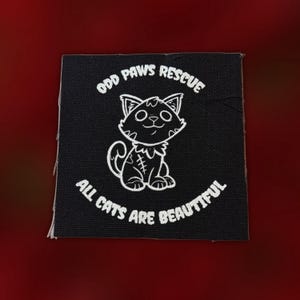 May include: A square black fabric patch featuring a white line art illustration of a cat with a stitched mark on its side. The text ODD PAWS RESCUE is printed in an arc above the cat, and ALL CATS ARE BEAUTIFUL is printed in an arc below the cat.