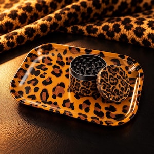 May include: A rectangular rolling tray and a cylindrical herb grinder, both featuring a brown and black leopard print pattern. The grinder is open, revealing black interior teeth, with its matching leopard print lid resting beside it.