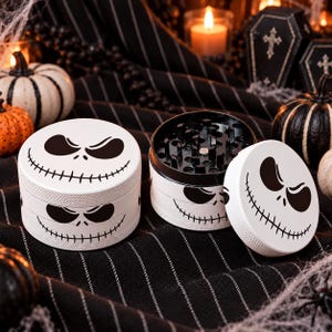 May include: Two cylindrical herb grinders with a white exterior featuring a black printed skeletal face design. One grinder is closed, while the other is open, revealing a black interior with sharp metal teeth and a mesh screen for processing herbs.