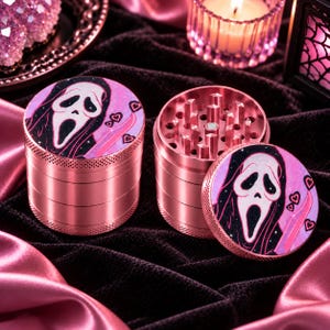 May include: Two metallic pink herb grinders with a four-piece design. The lids feature a graphic print of a white ghost-like mask with a black hood, a pink knife, and small pink hearts against a dark purple and pink starry background.