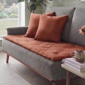 Puede incluir: Un sofá gris tapizado con un cojín de asiento capitoné de color óxido y dos cojines decorativos a juego. Una mesa auxiliar clara sostiene una pila de libros, una taza de cerámica y un jarrón de cristal con flores de color melocotón claro.