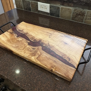 May include: A rectangular serving tray made of light-colored wood with a prominent, dark, irregular resin inlay running through the center. The tray features two black metal handles attached to the sides and has a smooth, polished finish.