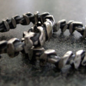 Op de afbeelding: Twee zilveren ringen met een textuurrijk, organisch ontwerp. De ringen hebben een reeks onregelmatige, rechthoekige vormen die met elkaar verbonden zijn, waardoor een unieke, industriële uitstraling ontstaat. De ringen worden gepresenteerd op een donkere, getextureerde ondergrond.