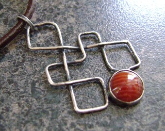 May include: A silver pendant necklace with a geometric design and a round, reddish-orange stone. The pendant is attached to a brown leather cord. The necklace is displayed on a dark, textured surface.