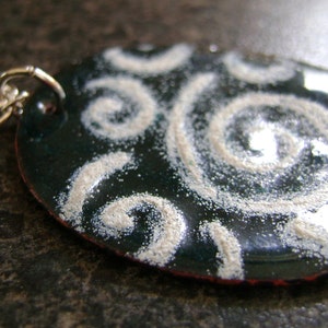 May include: Oval pendant necklace with a dark teal enamel finish and a silver-colored chain. The pendant features a white, swirling design. The necklace is a piece of jewelry.