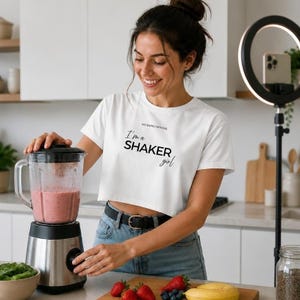 Op de afbeelding: Een wit kort T-shirt met korte mouwen en de zwarte tekst "NO EXPECTATIONS I'm a SHAKER girl" op de voorzijde. Het kledingstuk is gemaakt van een lichtgewicht stof en is afgewerkt met een ronde halslijn en een korte zoom die op de taille valt.