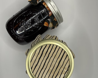 以下が含まれることがあります： 金属製の蓋が付いた2つのガラス製メイソンジャー。1つは暗い液体が入っており、カラフルな模様の黒い布で覆われています。もう1つは、細い黒の縦縞模様が入ったベージュの布で覆われています。