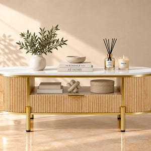 May include: An oval coffee table with a white marble top, a light wood fluted base, and gold metal legs. The table features a central drawer with a gold handle and an open shelf containing books, a decorative knot object, and a round box.