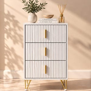 May include: A white three-drawer chest featuring a vertical ribbed texture on the drawer fronts, gold rectangular handles, and gold metal legs. The top surface holds a white vase with greenery, a wooden bowl, two books, and a glass reed diffuser.