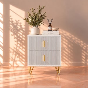 Puede incluir: Una mesita de noche blanca de dos cajones con textura acanalada vertical en los frentes, tiradores verticales dorados y cuatro patas de horquilla de metal dorado. Encima hay una planta verde, un difusor de aromas y dos libros apilados.