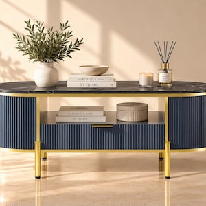 May include: An oval coffee table with a black marble top, navy blue fluted cabinet doors, and gold metal trim. The table features a central drawer with a gold handle, an open storage shelf, and four gold legs. It is styled with books and decor.