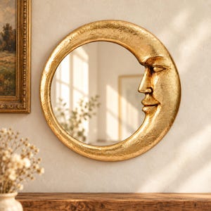 May include: A circular wall mirror featuring a gold-colored frame shaped like a crescent moon. The frame has a textured, metallic finish and incorporates a sculpted human face profile on the right side, with closed eyes and a gentle smile.