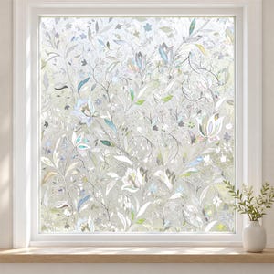 May include: A window film featuring a translucent, frosted finish with an intricate, repeating pattern of white and iridescent floral motifs, including stylised leaves, vines, and blossoms that catch the light to create a subtle, shimmering effect.