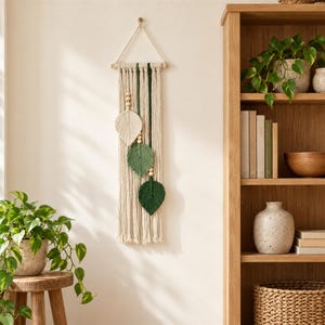 May include: A macrame wall hanging featuring cream-coloured cotton cord, a wooden dowel, and three leaf-shaped accents in cream, light green, and dark green. The piece is decorated with natural wooden beads and hangs from a simple cord loop.
