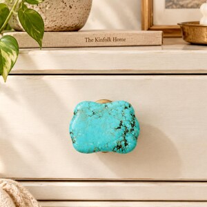 May include: A cabinet knob featuring a polished, irregular, rectangular-shaped piece of bright turquoise stone with dark brown matrix veining, mounted on a circular gold-toned metal base attached to a light-colored wooden drawer front.