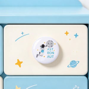 May include: A round white drawer knob featuring a graphic of an astronaut reaching for a gray moon, surrounded by yellow stars and the text AST RON AUT, mounted on a cream-colored drawer front within a light blue wooden furniture frame.