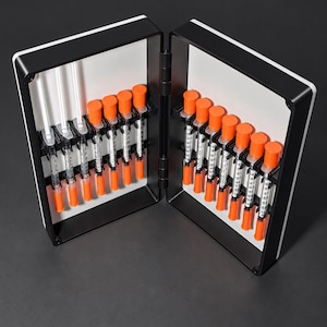 May include: A black and white hinged storage case containing a series of clear plastic syringes with bright orange caps. The syringes are arranged in vertical slots within the interior of the case, featuring clear measurement markings on each barrel.