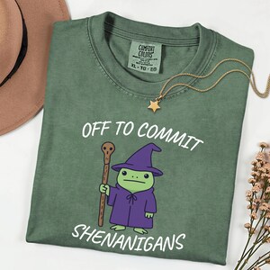 Könnte beinhalten: Ein salbeigrünes T-Shirt mit einer Grafik eines grünen Frosches, der einen violetten Zaubererhut und eine Robe trägt und einen Holzstab hält. Der Text "OFF TO COMMIT SHENANIGANS" ist in Weiß über und unter der Froschgrafik aufgedruckt.