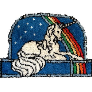 May include: A whimsical, rectangular rug featuring a white unicorn with black accents, a rainbow, and a blue sky with white stars. The rug has a textured, tufted appearance and a blue border.